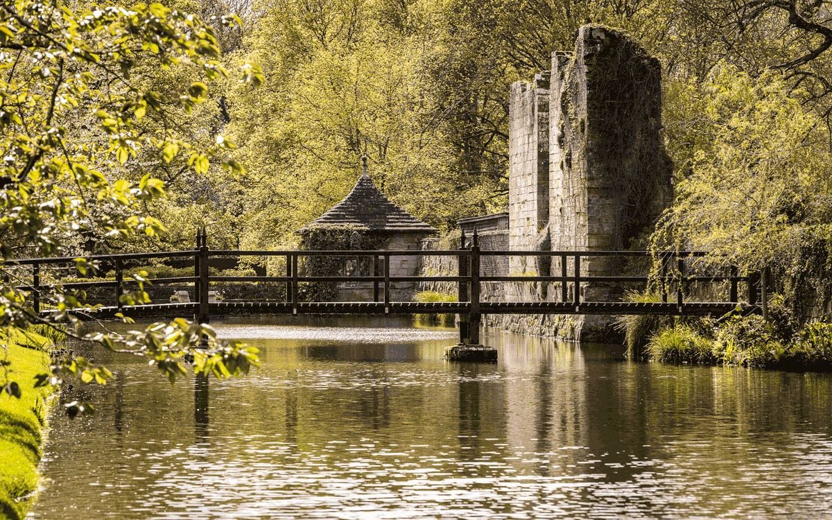 A wooden footbridge over the moat, with the ruined manor tower behind the trees
